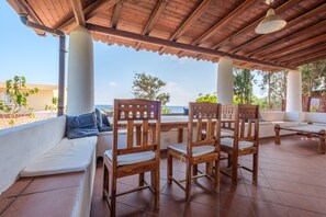 Un espacio de estar al aire libre techado, con sillas y sofá de madera, piso de baldosas y vista al mar.