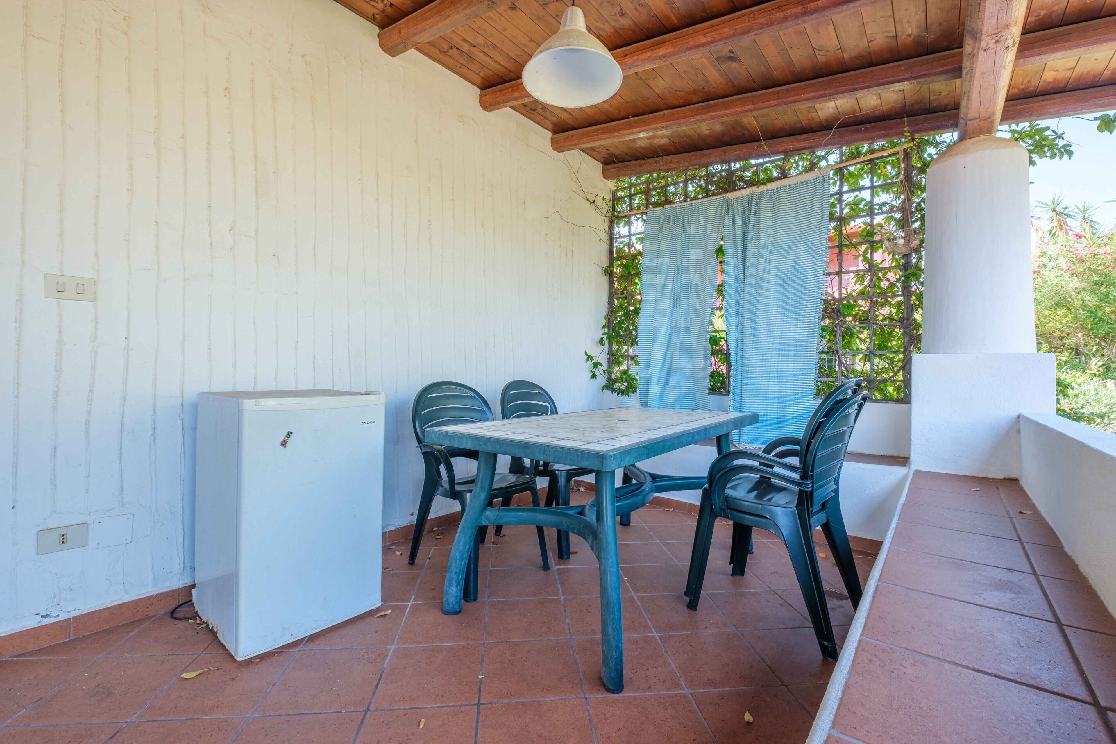 Overdekt terras met een tafel en stoelen, een kleine koelkast en uitzicht op groen door een raam.