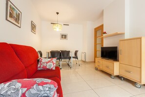 Una sala de estar con un sofá rojo, un televisor sobre un mueble de madera, una mesa de comedor con sillas e iluminación en el techo.