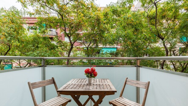 Ein Balkon mit Holztisch und -stühlen, einem Blumentopf mit roten Blüten und Blick auf Gebäude und Bäume.