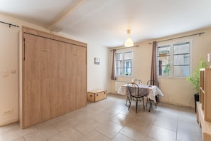Ein Zimmer mit Holzschrank, Tisch mit Stühlen, einer Pendelleuchte und Fenstern mit Vorhängen.
