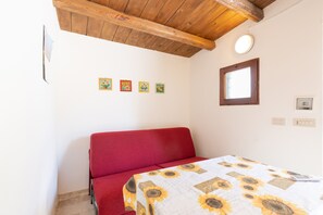 A room with a red sofa, a bed with a sunflower-patterned bedspread, wooden ceiling beams, and a small window.