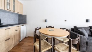 A compact kitchen with wooden cabinets, a white dishwasher, a round wooden table with chairs, and a dark grey sofa.