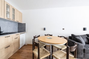 Une cuisine compacte avec des meubles en bois, un lave-vaisselle blanc, une table ronde en bois avec des chaises et un canapé gris foncé.