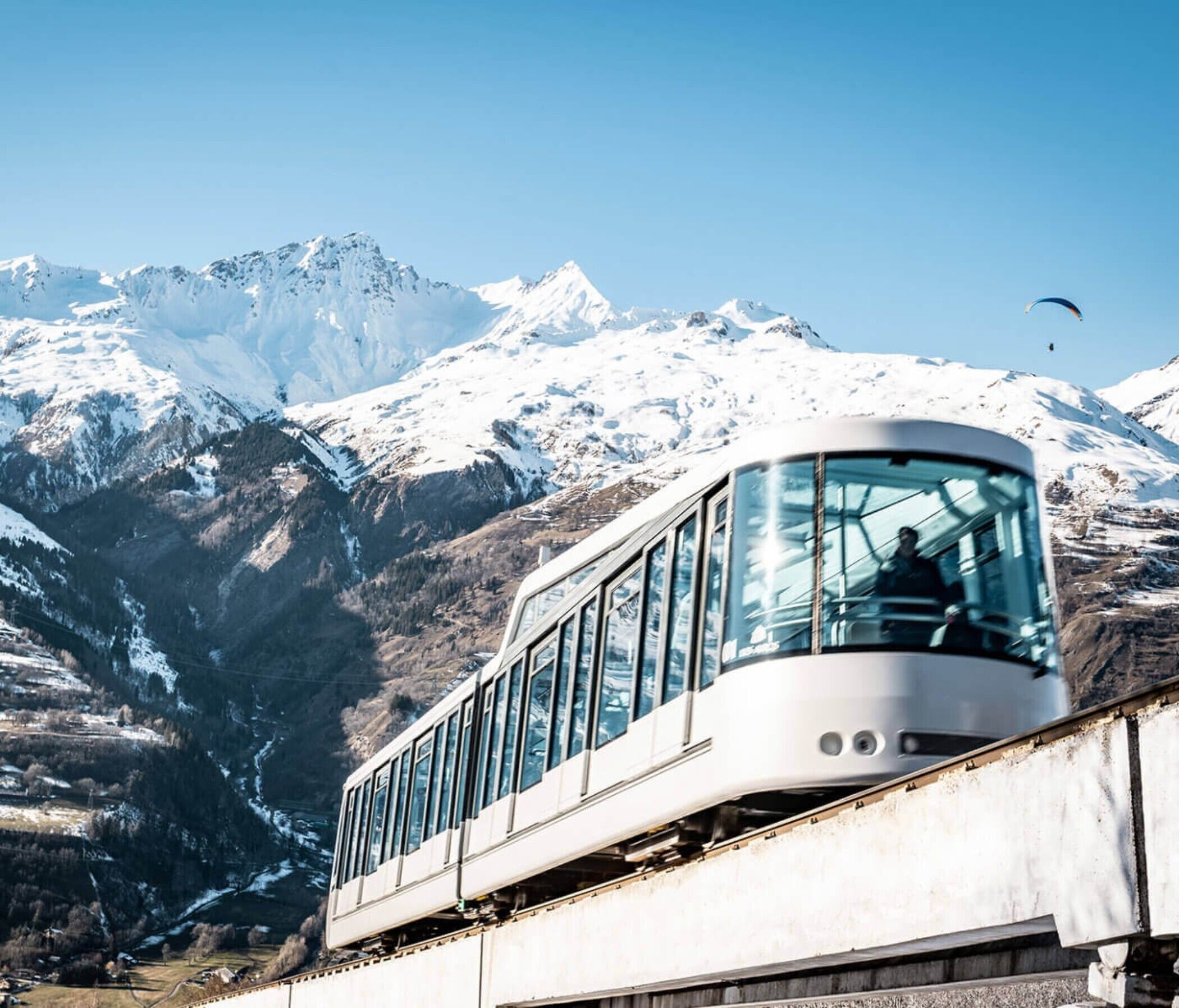 Un train moderne aux grandes fenêtres, circulant sur une voie ferrée avec, en arrière-plan, des montagnes enneigées.