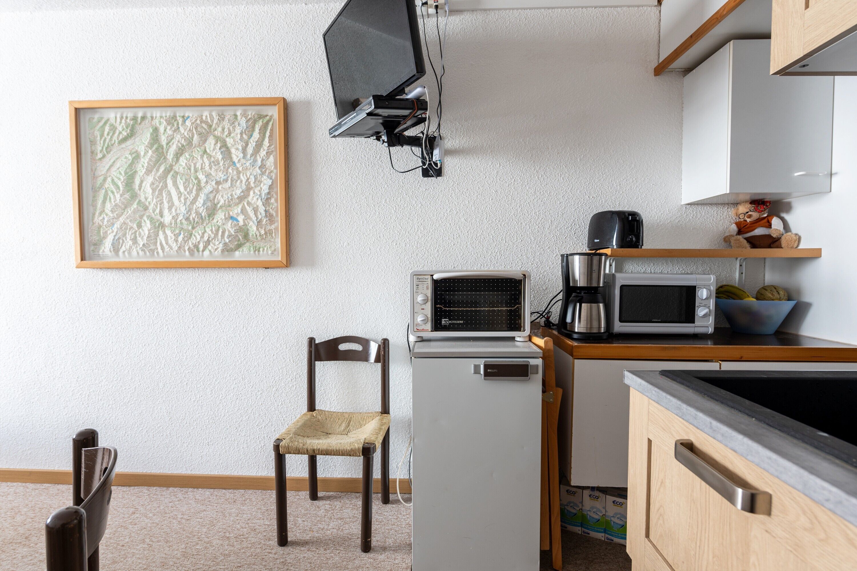 A compact kitchen with a microwave, toaster, and a small refrigerator. A chair is placed against the wall, and a framed picture hangs above the counter.