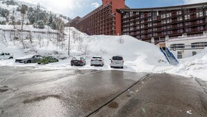 A multi-story building with a red facade, parking area with snow-covered cars, and a snowy landscape.