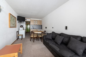A compact living space with a sofa, a dining table, chairs, a small kitchen area, and a television mounted on the wall.