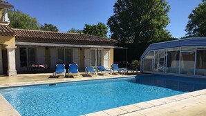 A swimming pool with blue water, surrounded by beige tiles. There are blue lounge chairs and a covered area with glass walls. - Holiday Home 'Maison Piscine Chauffe Jacuzzi' with Private Terrace, Private Garden and Wi-Fi (Tourrettes)