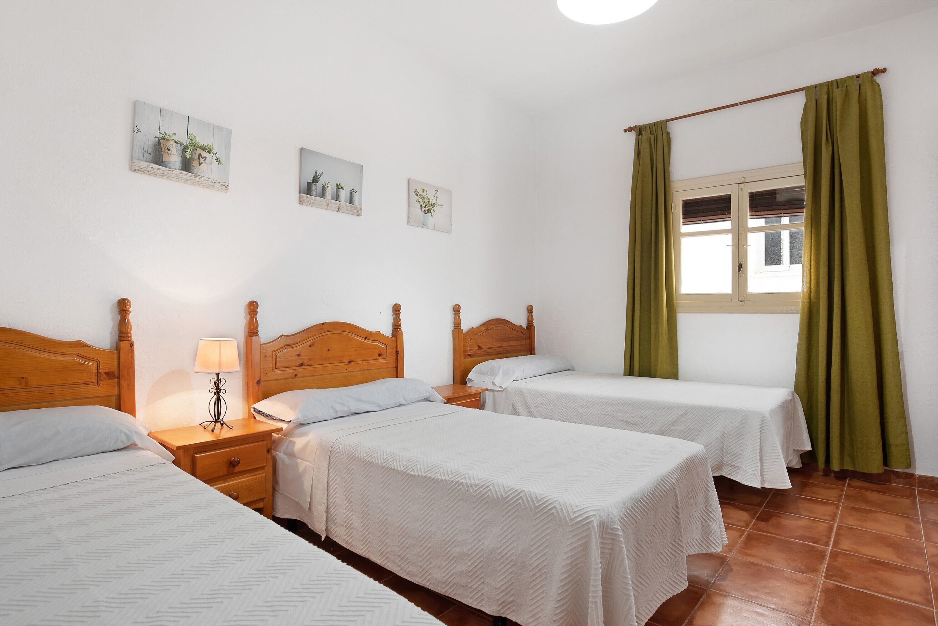 Une chambre avec trois lits, des têtes de lit en bois, une table de chevet avec une lampe et une fenêtre avec des rideaux.