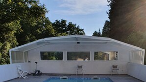 A swimming pool with a hot tub, surrounded by a covered patio with chairs and a window overlooking the pool.
