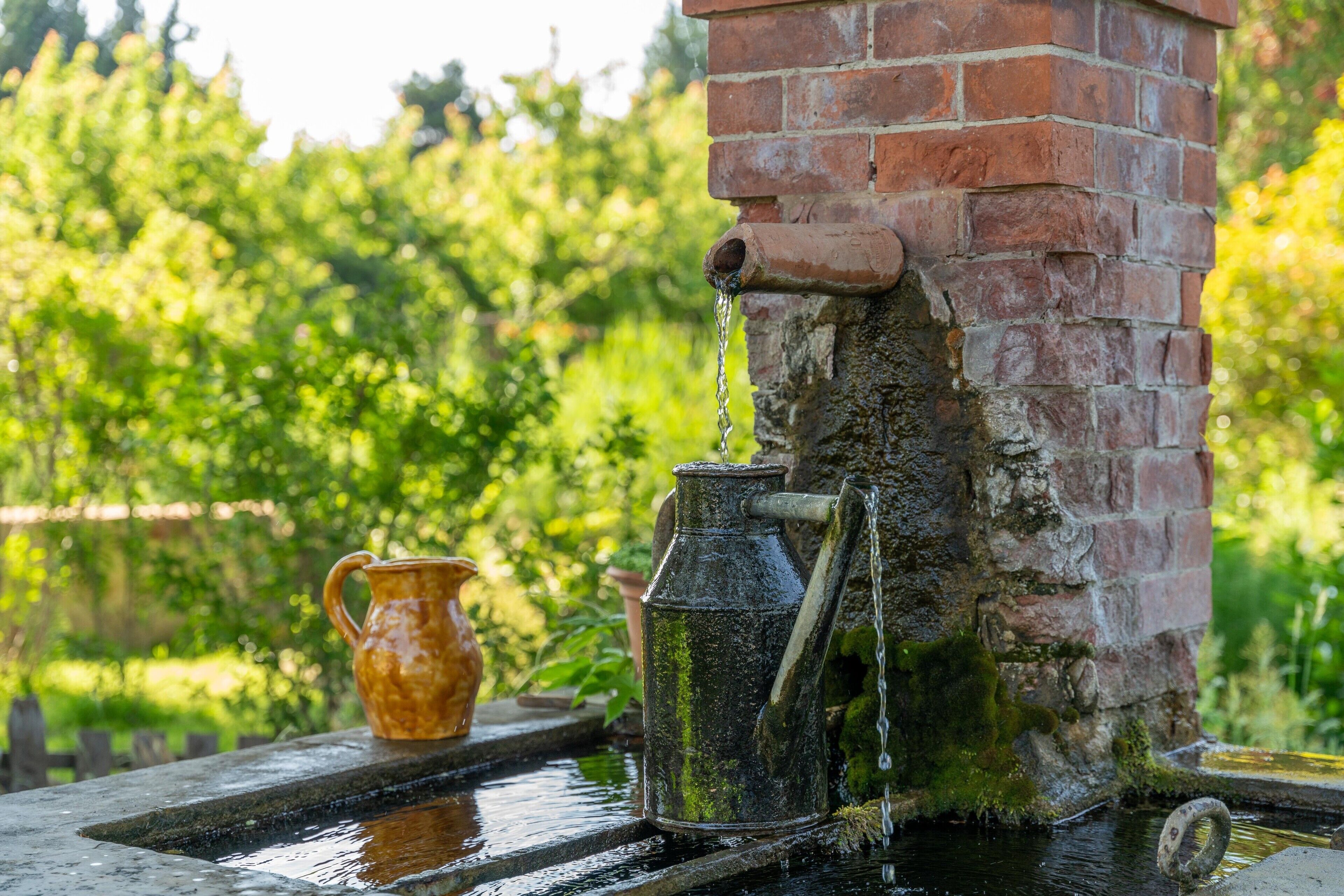 A rustic outdoor water feature with a brick wall, a metal spout, and a metal jug.