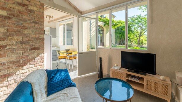 A modern living room with a brick wall, a glass table, a sofa with blue cushions, a TV on a wooden stand, and a large window with a view of greenery.