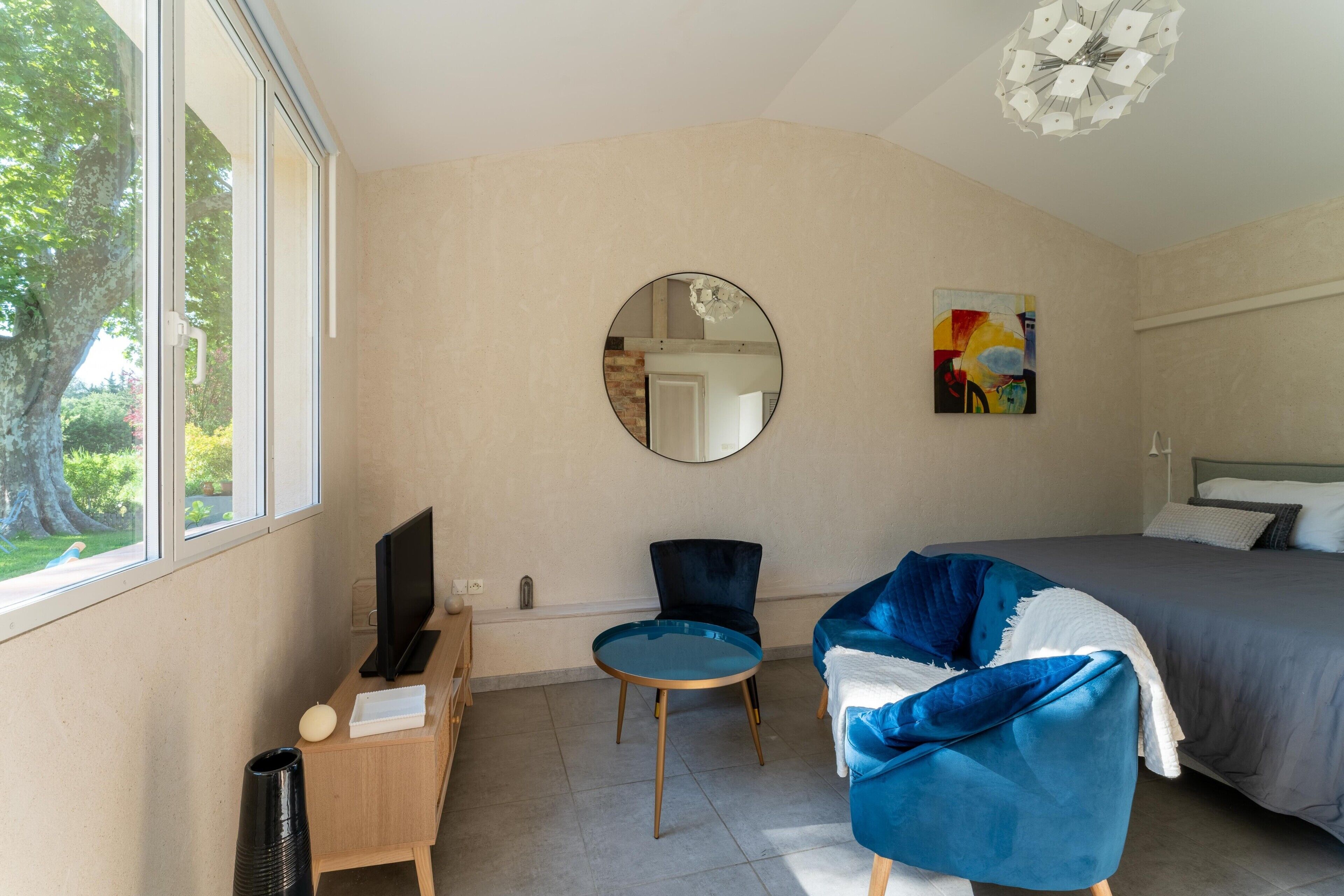 Un dormitorio con cama, un espejo redondo, una silla, una mesita, un televisor y una ventana.