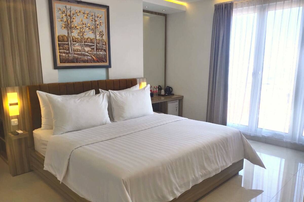 A hotel room with a large bed, a nightstand, a desk with a lamp, a painting on the wall, and a window with curtains.