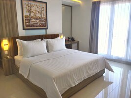 A hotel room with a large bed, a nightstand, a desk with a lamp, a painting on the wall, and a window with curtains.