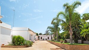 A courtyard with a tiled pathway, palm trees, and a white building with a tiled roof. - Holiday Home "Chalet Fuente3 Shared Pool Family" with Shared Pool and Garden (Conil de la Frontera)