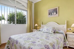 A bedroom with a bed, a nightstand, a lamp, a window with curtains, and a painting on the wall.