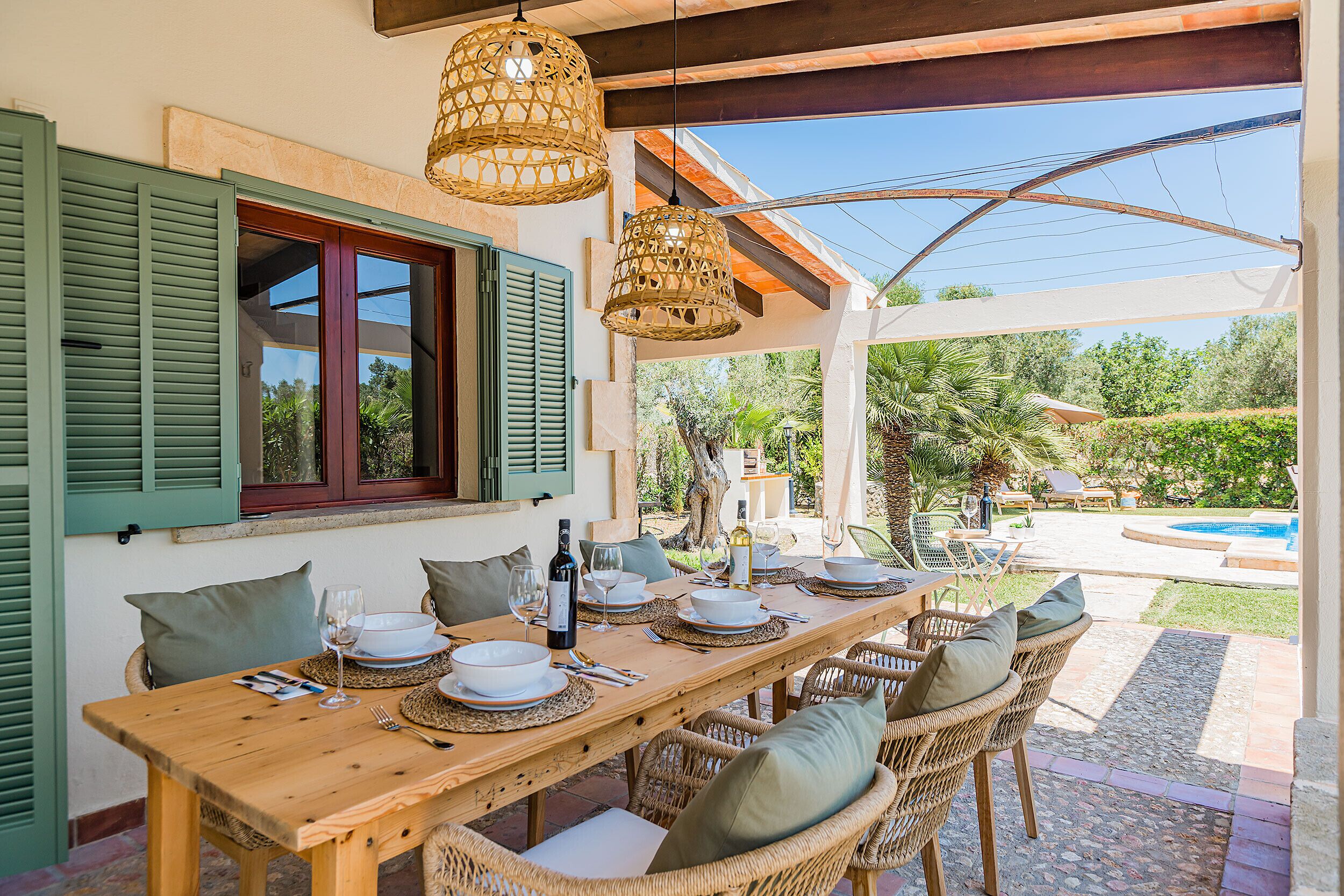 An outdoor dining area with a wooden table set for a meal, wicker chairs, and a view of a swimming pool.