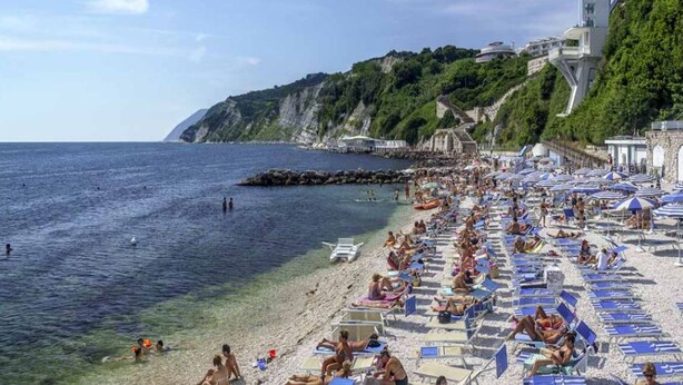 A crowded beach with numerous sun loungers and umbrellas, a rocky coastline, and a large building with a unique architectural design. - Apartment in Ancona near Passetto Beach (Ancona)
