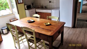 Una mesa de madera con sillas, una cocina con microondas, refrigerador y ventana con vista a follaje.