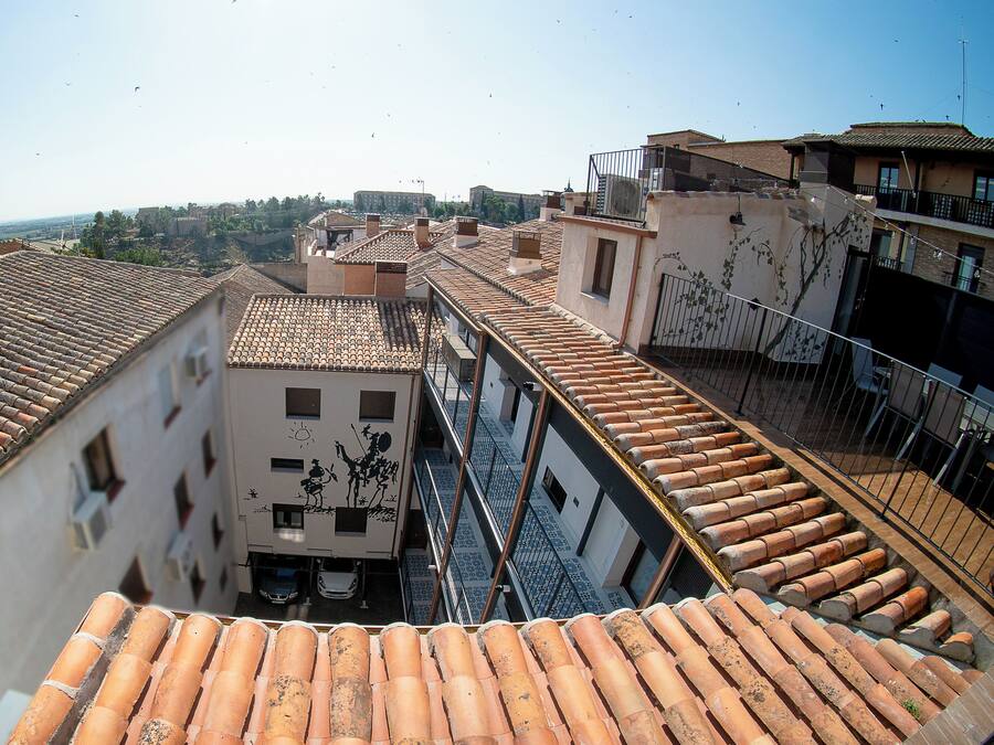 Vista de techos de tejas de terracota y edificios con balcones, cielo despejado y colinas a lo lejos.