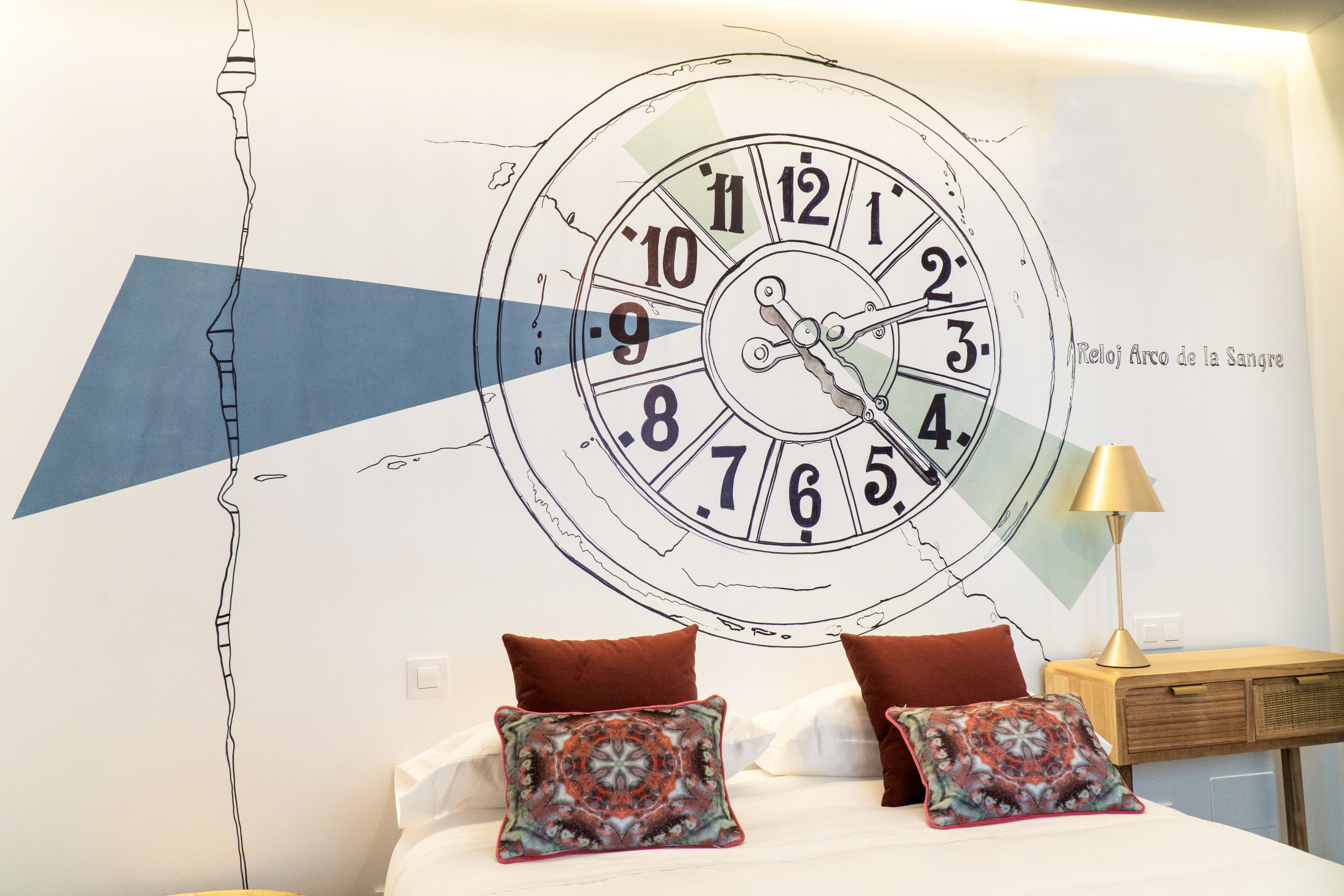 Un dormitorio con un reloj de pared grande, dos almohadas, una mesita de noche y una lámpara.