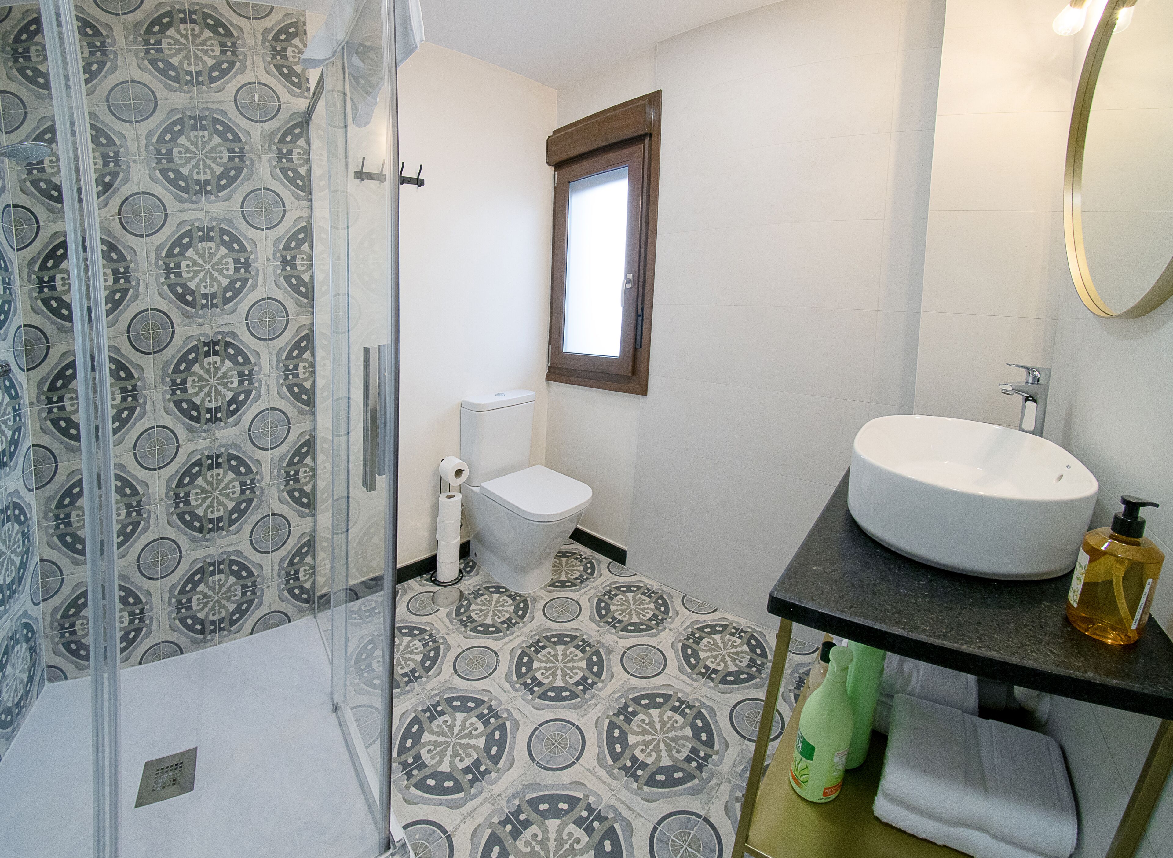 Un baño con baldosas estampadas en el suelo, un inodoro, un lavamanos con un espejo encima y una zona de ducha.