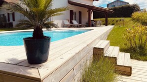 Un espace aménagé au bord de la piscine, avec une terrasse en bois, un palmier et une maison au toit rouge.
