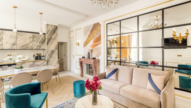 A modern living room with a beige sofa, teal armchairs, and a coffee table. A large mirror and chandelier are visible. - Merveil - Luxury Suite - Tour Eiffel - Bac I (Paris)