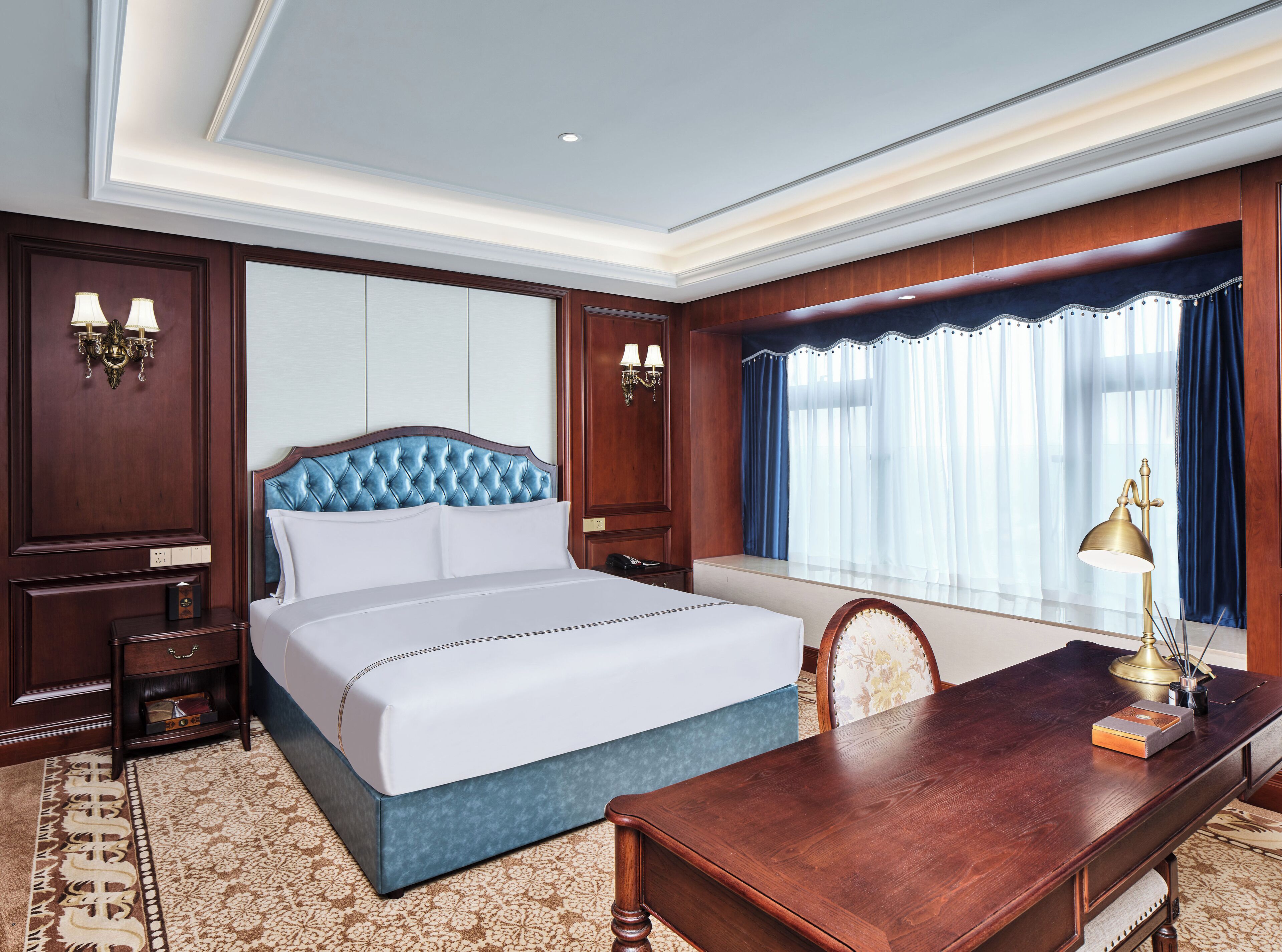 A hotel room with a large bed, a wooden desk, a nightstand, and a window with curtains.