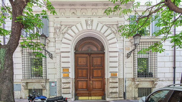 Une grande porte en bois richement décorée, surmontée d’une arche ornementale et flanquée de deux fenêtres avec des barreaux en métal.