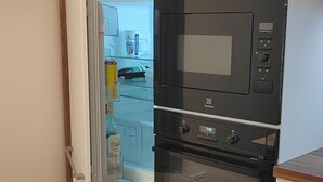 A compact kitchen with a built-in microwave and oven, a white refrigerator, and a wooden shelf above the fridge. - New apartment in the quiet of the Jizera Mountains (Smržovka)