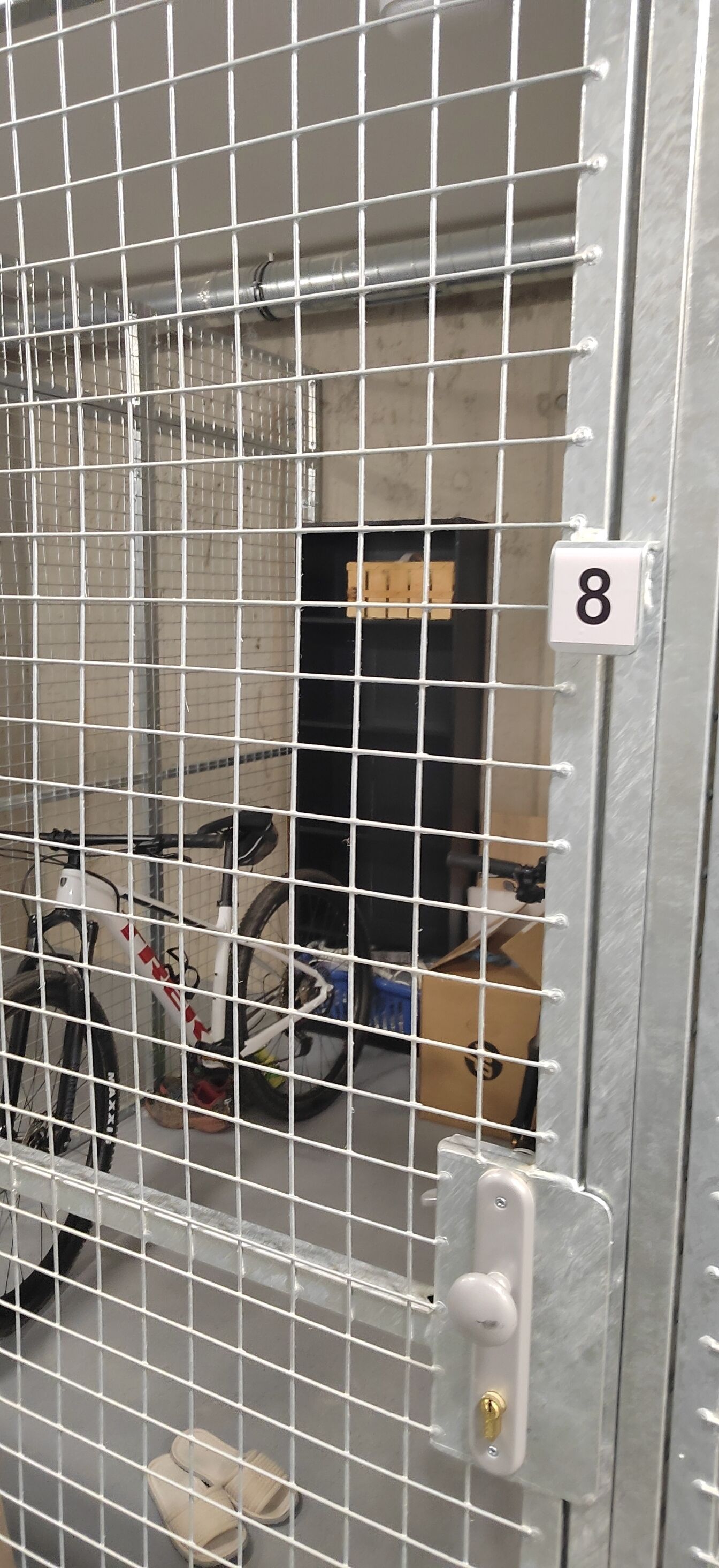 Une porte en métal sur laquelle est apposé un panneau portant le chiffre 8, un vélo et une paire de chaussons posés sur le sol.