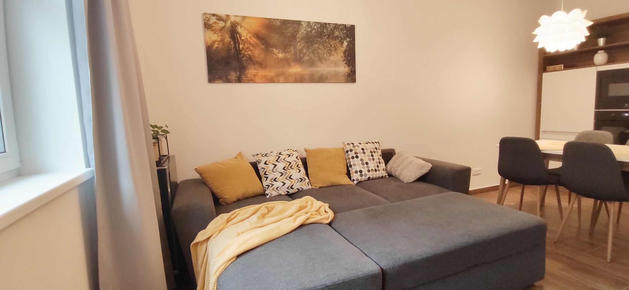 A modern living room with a grey sectional sofa, a dining table with chairs, a large abstract painting on the wall, and a pendant light.