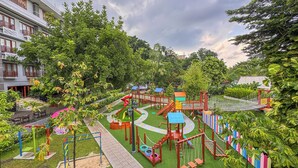 Ein Spielplatz mit verschiedenen Spielgeräten, umgeben von Grünflächen und einem Gebäude im Hintergrund.