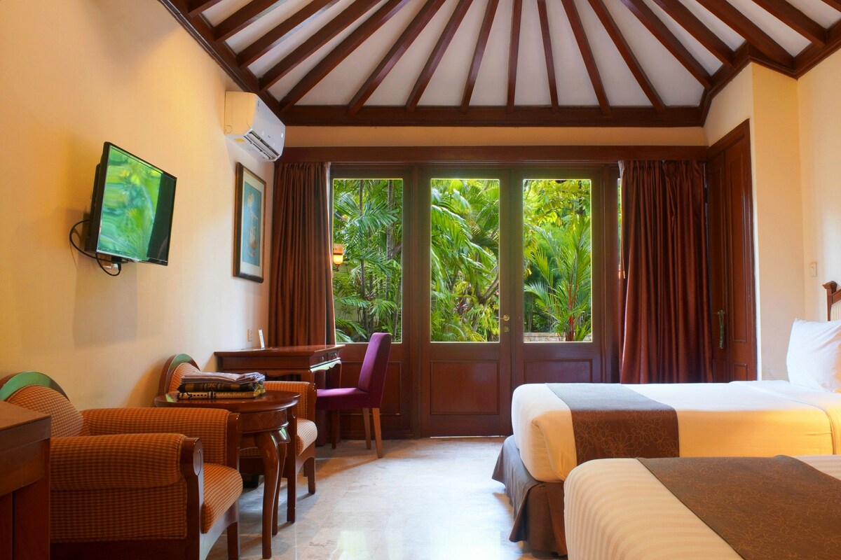 A hotel room with a bed, a desk with a chair, a television, and a view of lush greenery through the windows.
