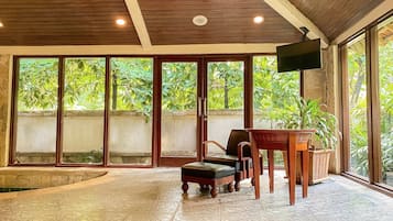 Una stanza con soffitto in legno, ampie porte scorrevoli in vetro, una televisione a schermo piatto, una panca di legno e un piccolo tavolo con una pianta.