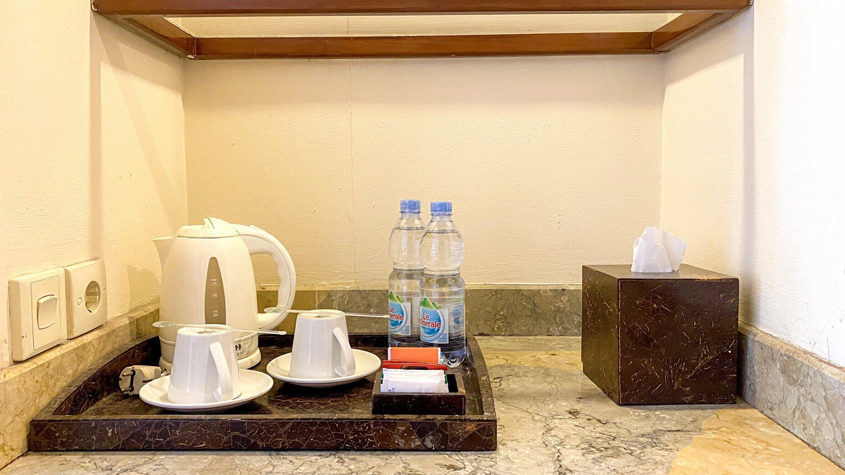 A hotel bathroom with a marble countertop, a coffee maker, two cups, two bottles of water, a tissue box, and a towel.