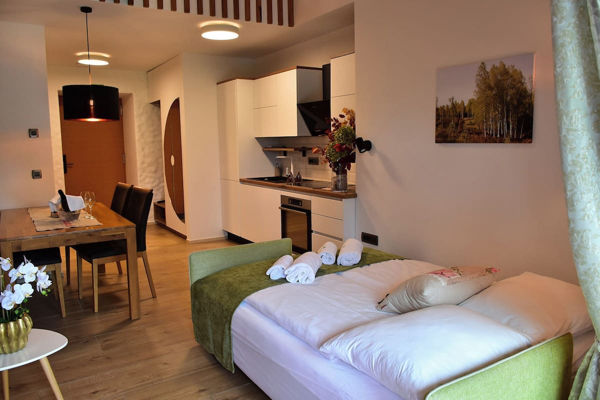 A modern hotel room with a bed, a dining table with chairs, a kitchen area, and a bathroom.