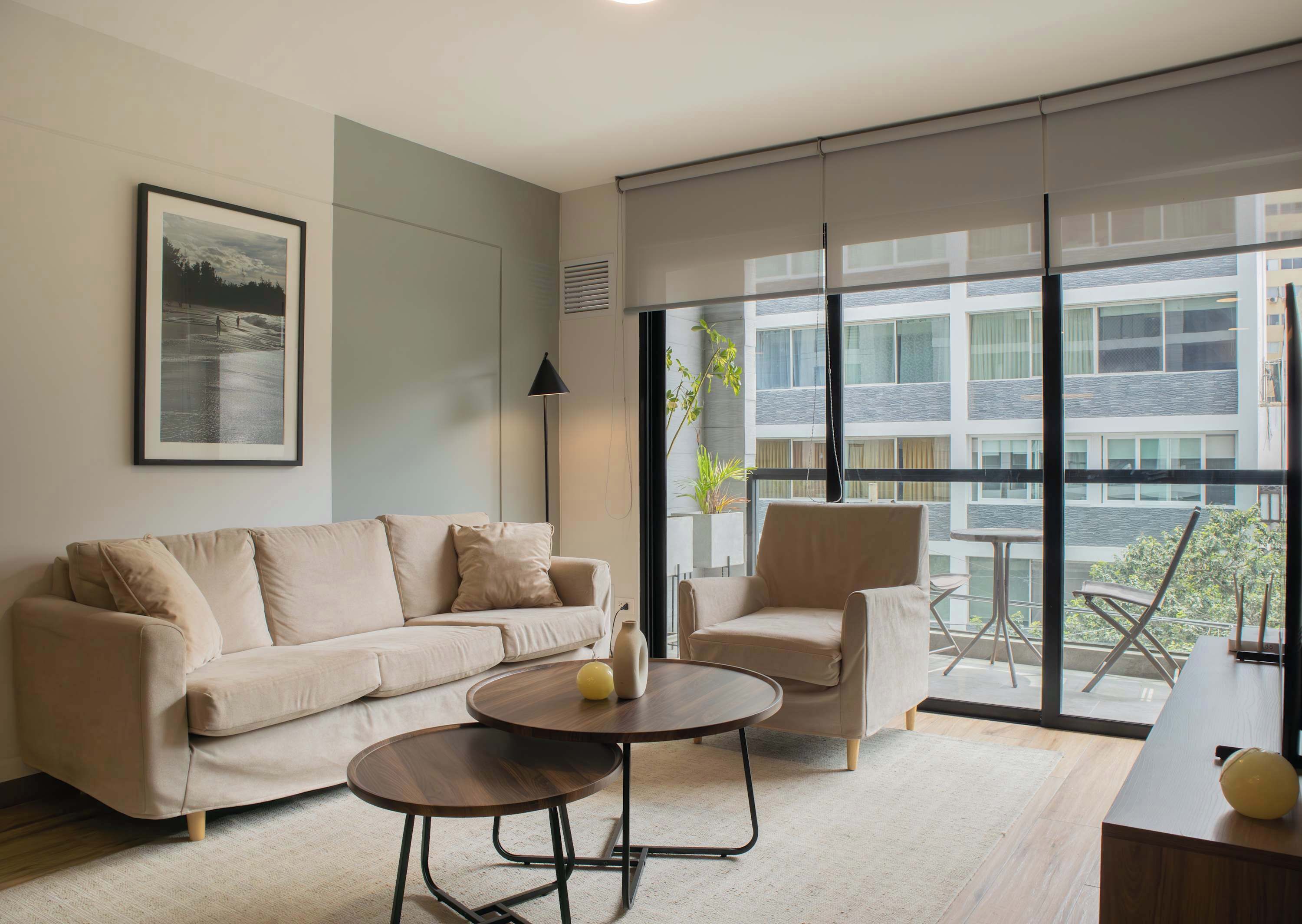 Ein modernes Wohnzimmer mit einem beigen Sofa, einem runden Couchtisch und einem Sessel. Es gibt ein großes Fenster mit Blick auf ein Gebäude und eine Pflanze auf dem Fensterbrett.
