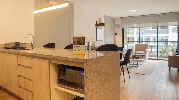 A modern kitchen with a built-in oven, a coffee machine, and a large island with a sink and a mirror. There is a dining area with a table and chairs, and a large window offering a view of the city.