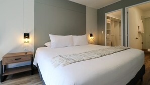 A neatly made bed with a white comforter and pillows, a bedside table with a lamp, and a wardrobe with a mirror.