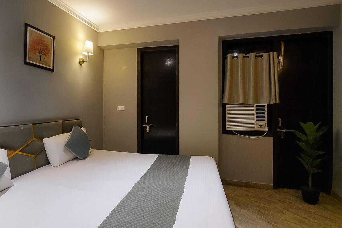 Ein Hotelzimmer mit Bett, Wandlampe, Tür, Fenster mit Vorhängen und einer Topfpflanze.