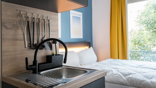 A modern kitchen with a built-in sink, utensils hanging on the wall, and a view of a bedroom with a bed and window. - Antares Apartments (Düsseldorf)