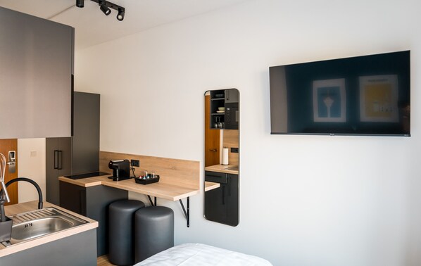 Una kitchenette compacta con fregadero, cocina y encimera, un pequeño rincón de descanso con taburetes, y un televisor de pantalla plana montado en la pared.