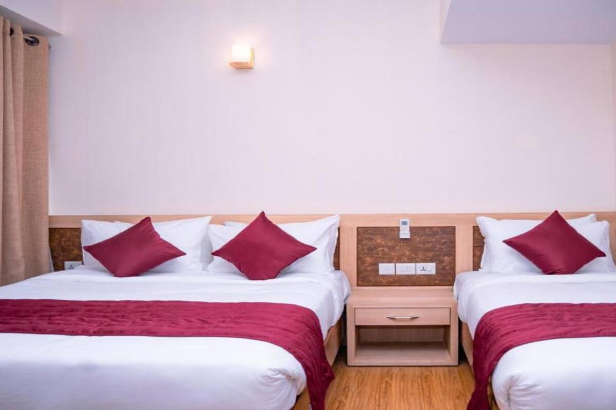 A hotel room with two beds, each with white linens and red pillows. There is a wooden nightstand between the beds, a wall-mounted air conditioner, and a small wall-mounted light.