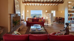 A spacious living room with a red sofa, a coffee table, and a fireplace. There is a bookshelf and a dining area visible in the background. - Villa Grene - Homelike Villas (Grosseto)