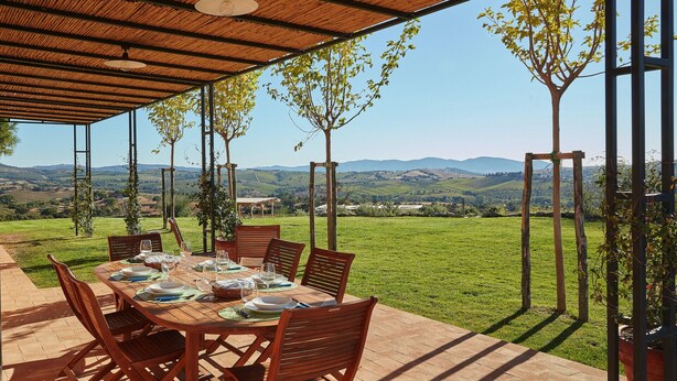An outdoor dining area with a table set for four, surrounded by wooden chairs, under a wooden pergola with a view of rolling hills and a clear sky. - Villa Grene - Homelike Villas (Grosseto)