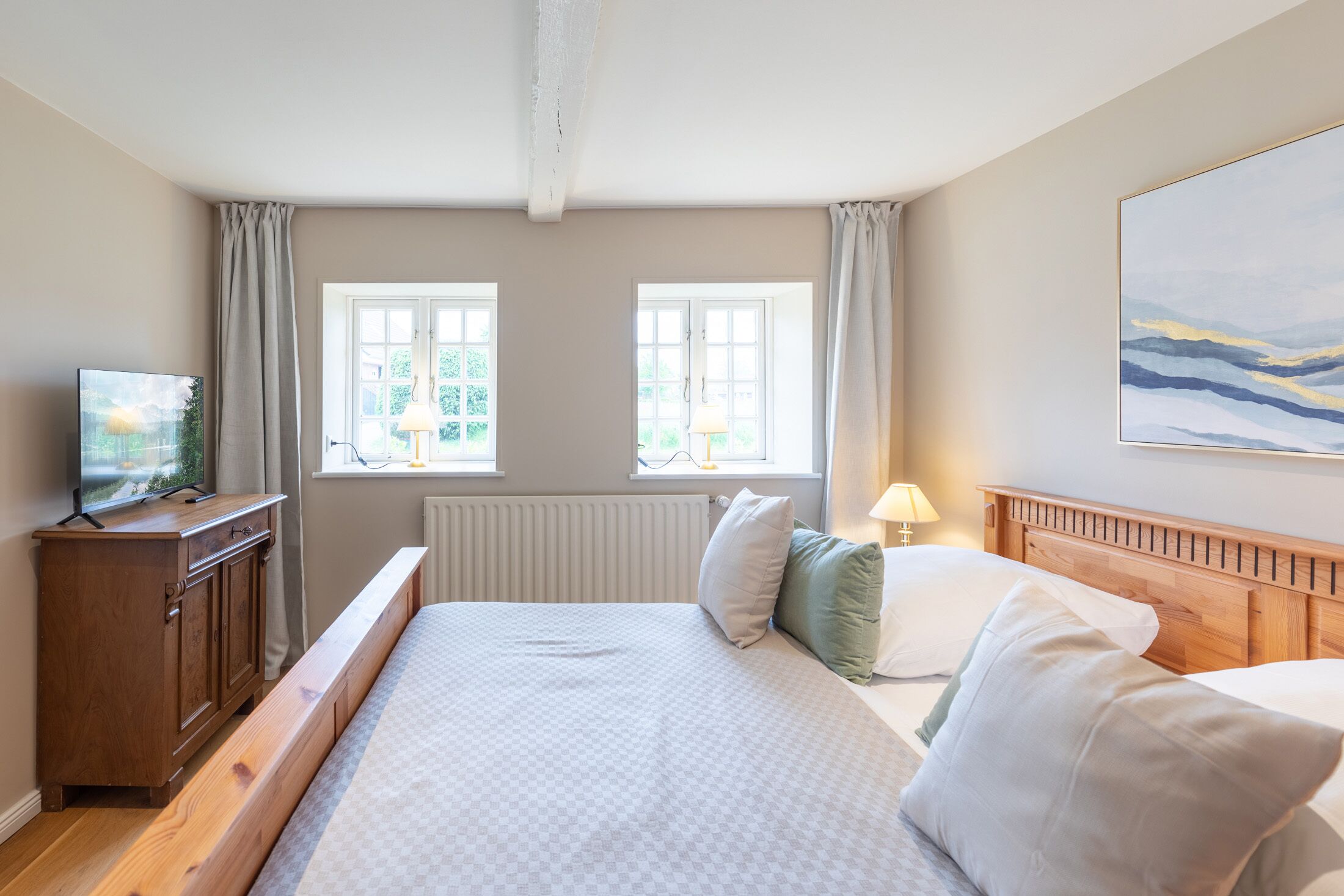 A bedroom with a bed, a wooden dresser, two windows, a radiator, and a painting on the wall.